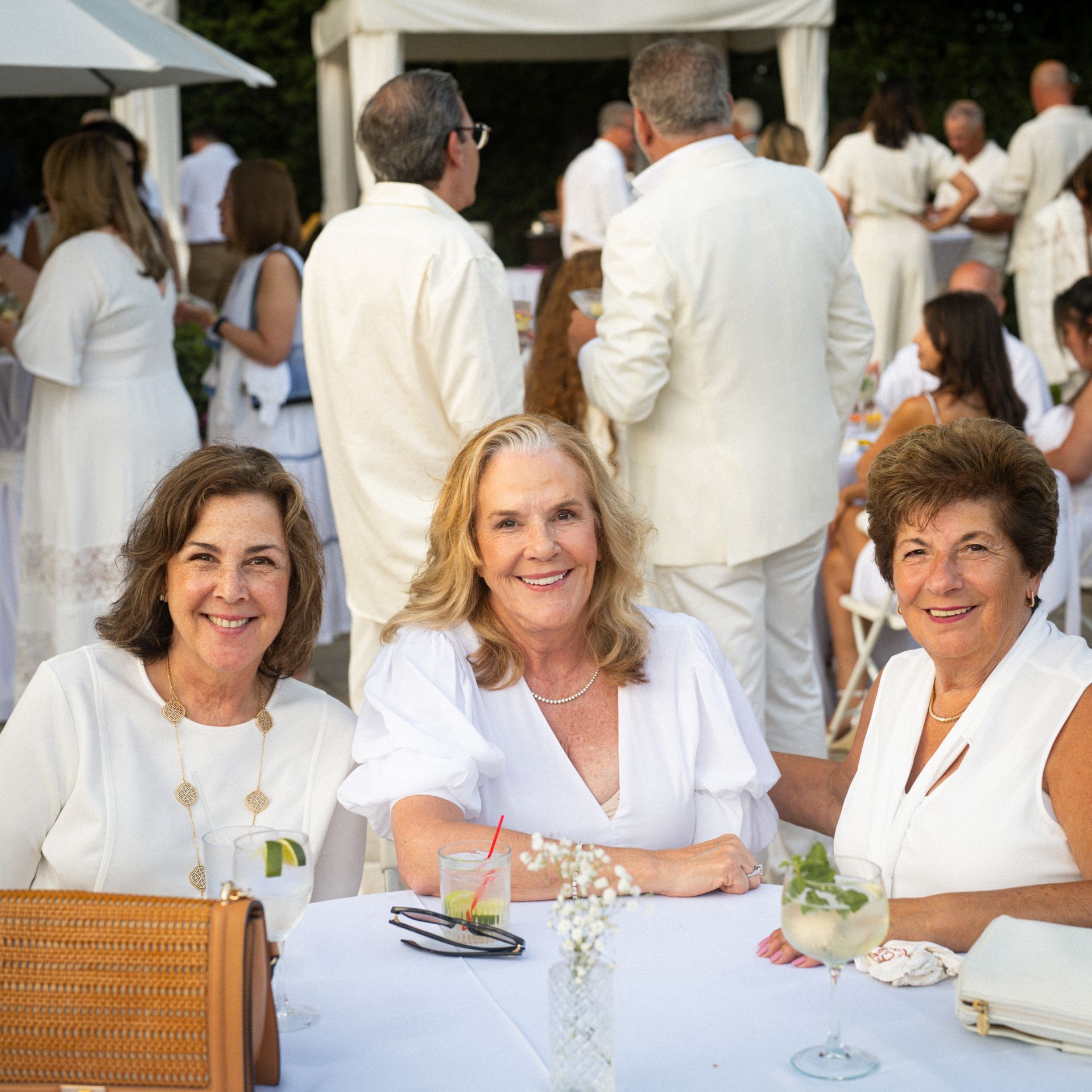 Three women in white clothing at an outdoor gathering, smiling at the camera. Others in white attire are in the background.