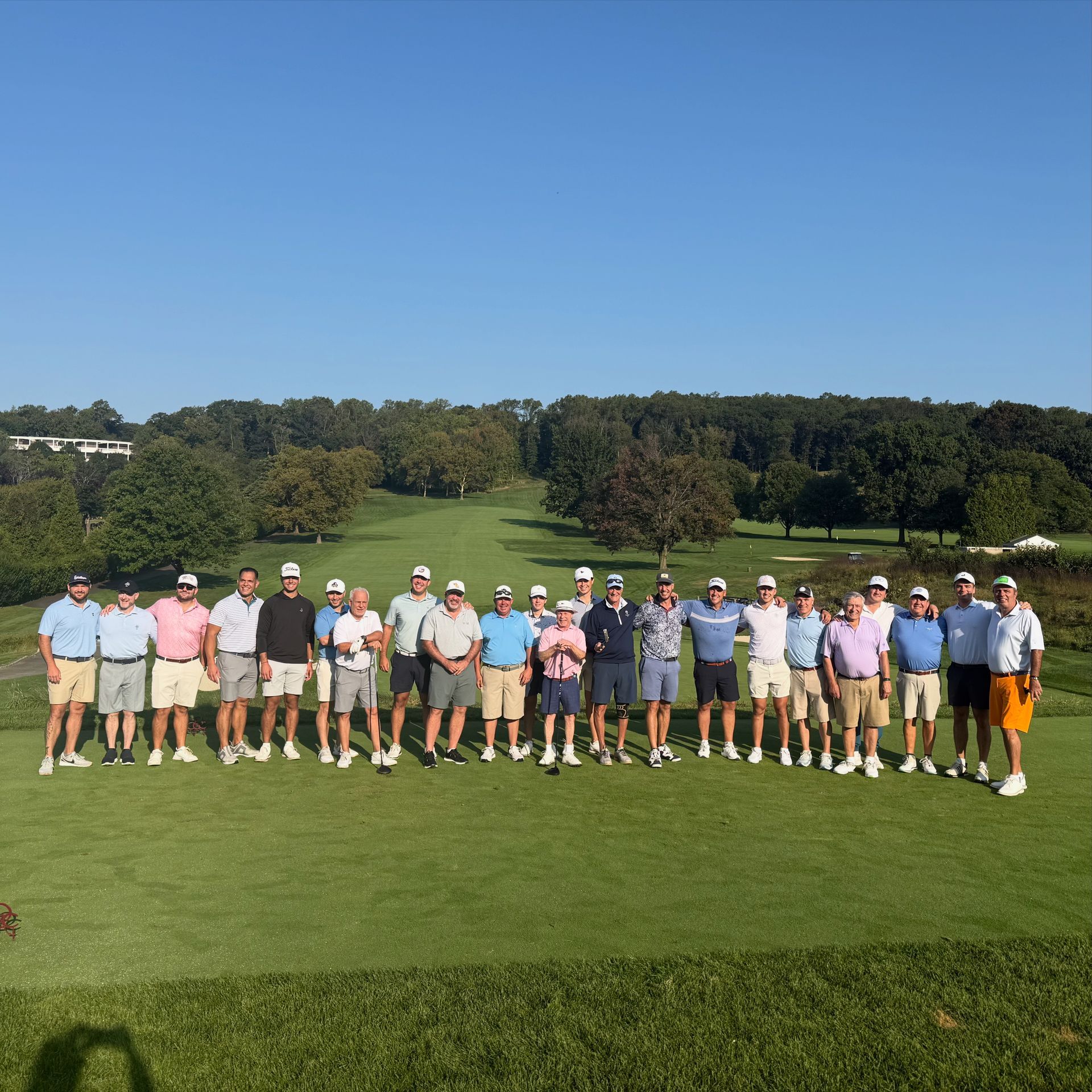 Group of golfers on a green, posing for a photo. They wear golf attire. Trees and a golf course are in the background.