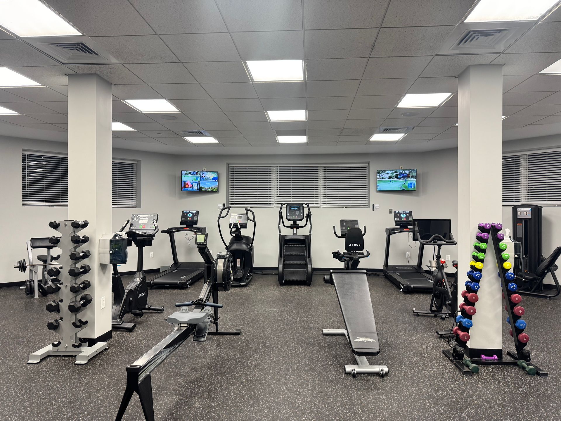 A well-lit gym with exercise equipment: treadmills, bikes, weights, and TVs, arranged on a black floor.
