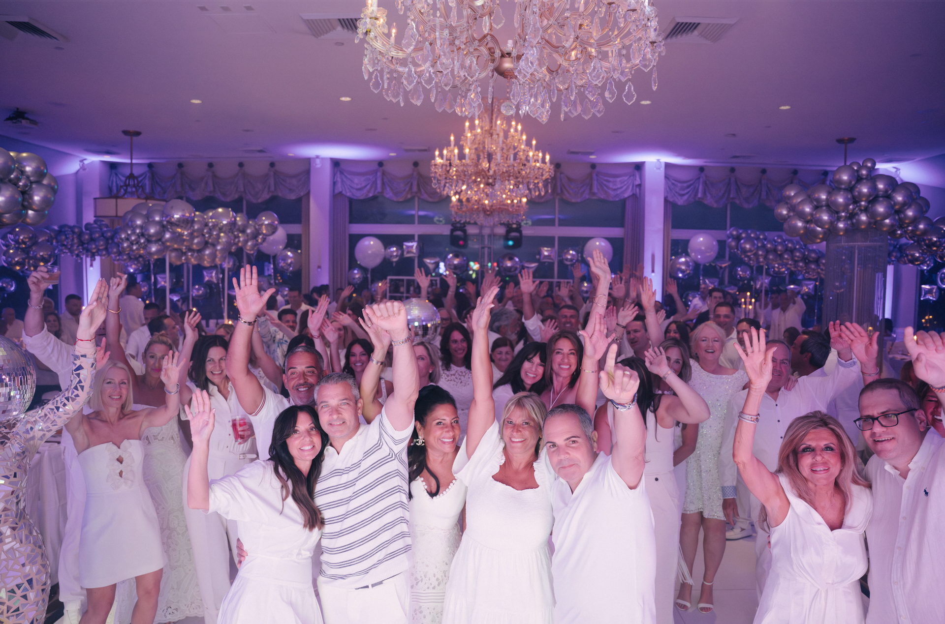 Large group of people in a white room celebrating, raising arms. Glittering chandelier and balloons overhead.