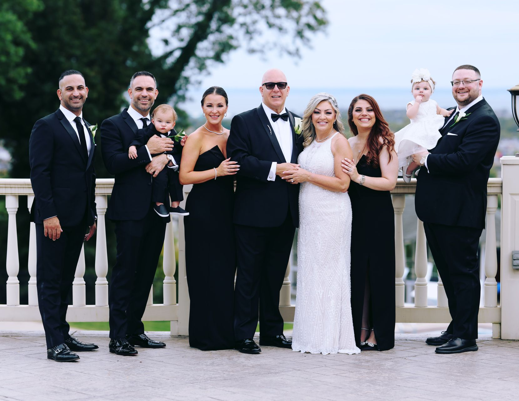 Group of people posing outdoors on a patio, wearing formal attire. Some hold children.