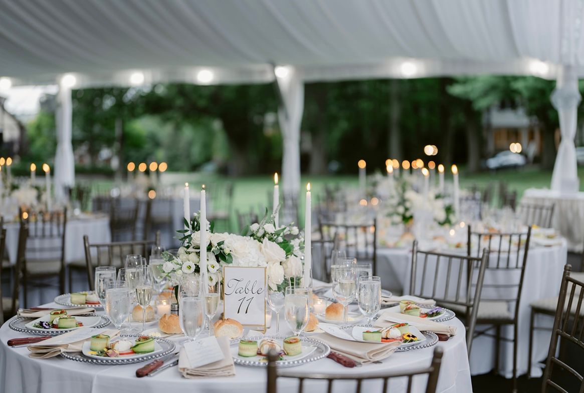 Elegant outdoor wedding reception with round tables set under a white tent, lit by candles and string lights.
