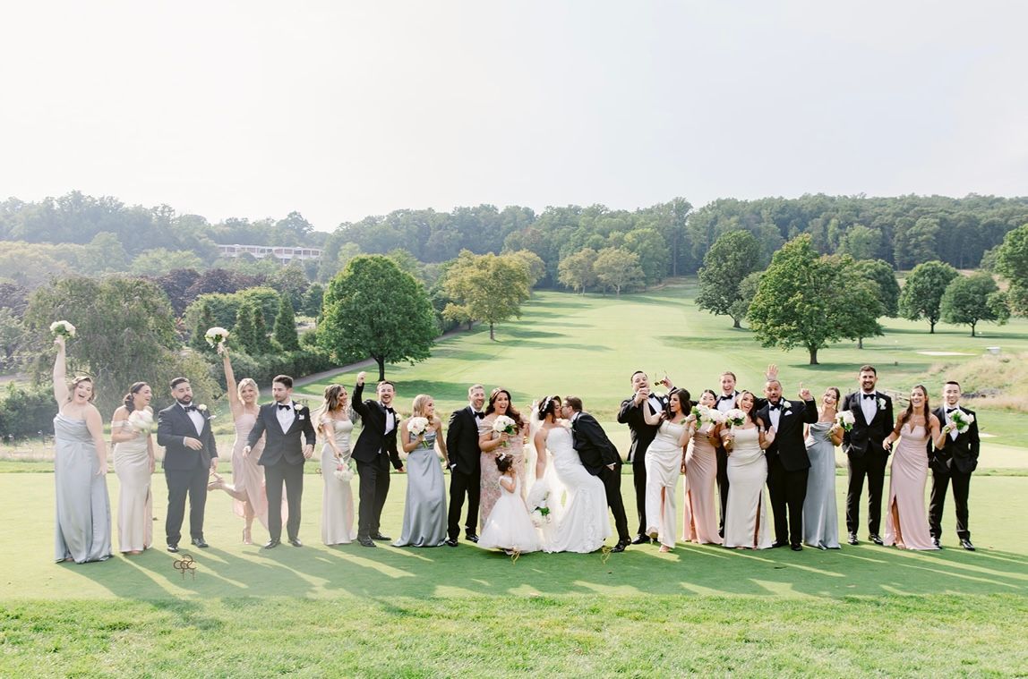 Wedding party celebrating on a golf course; the bride and groom kiss, guests raise arms. Green landscape, sunny day.