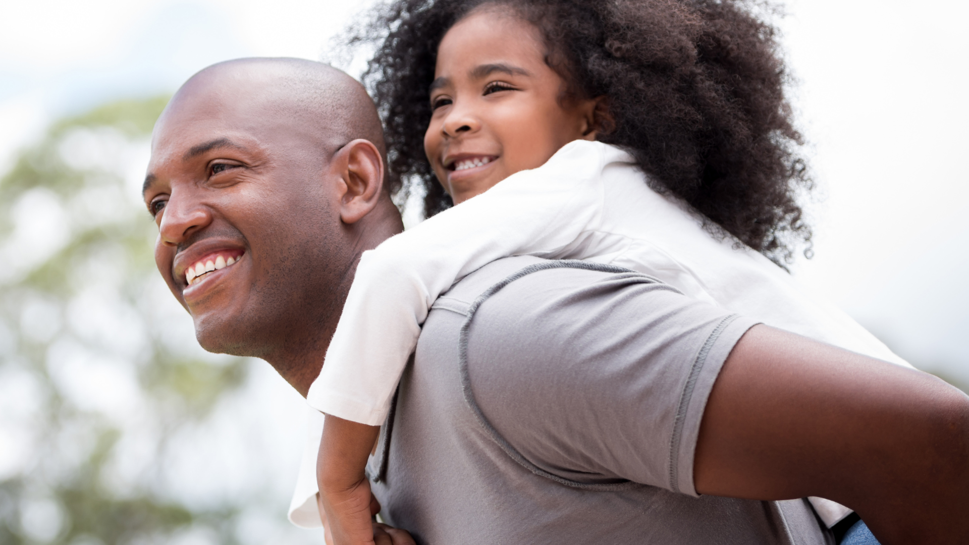 Smiling Black man giving a piggyback ride to a smiling Black girl outdoors.