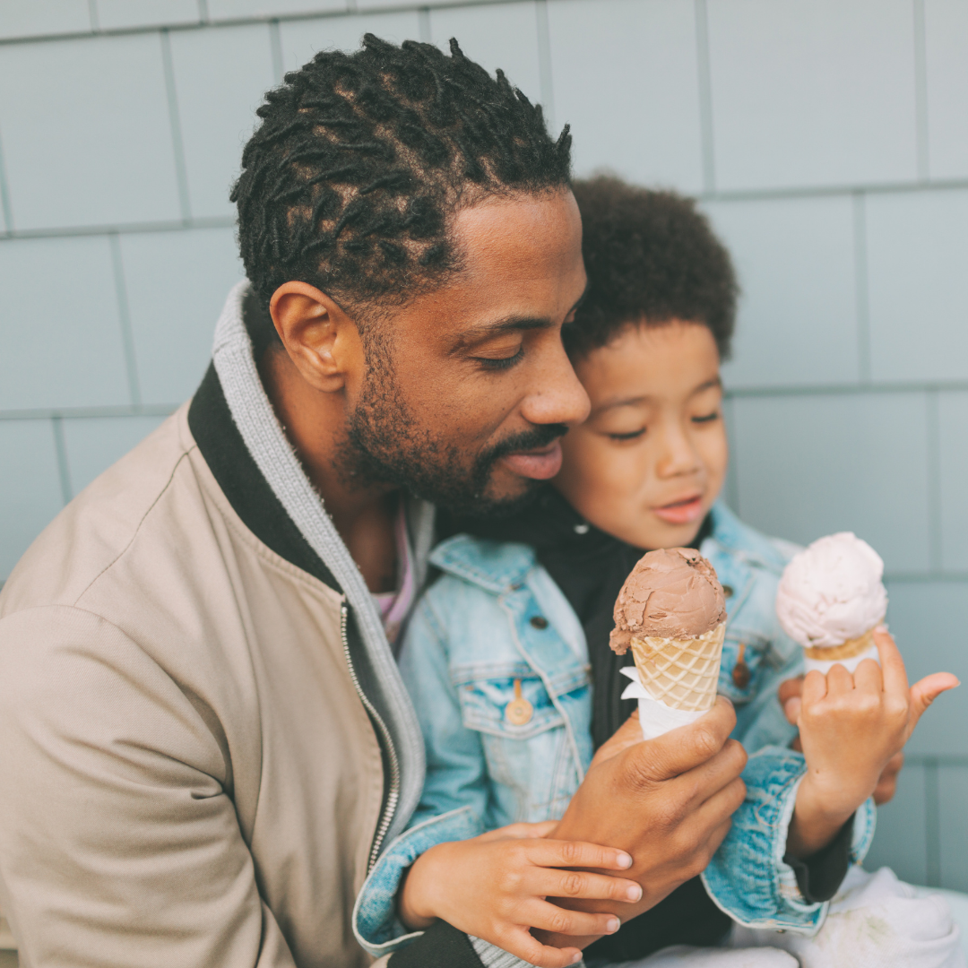 Father and son share ice cream cones outside; father smiles, son looks at ice cream.