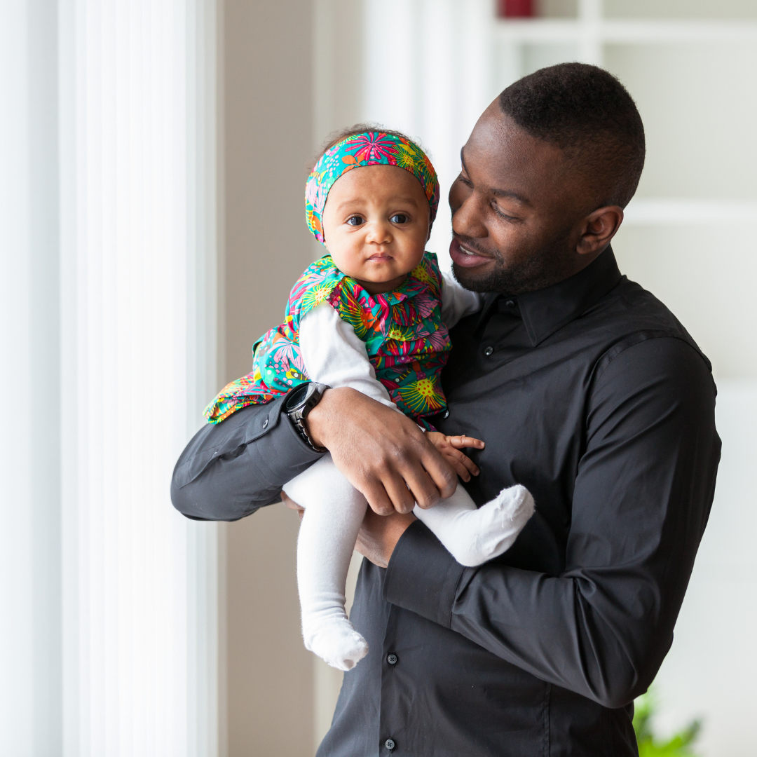 Black man holding a baby girl near a window. Both are looking at the camera.