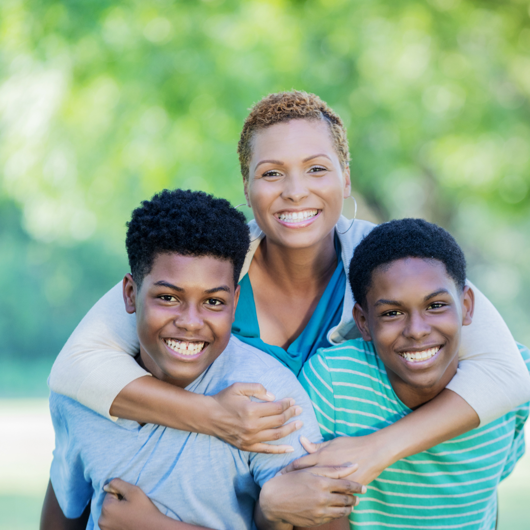 Woman smiling, hugging two teenage boys outdoors in a park.