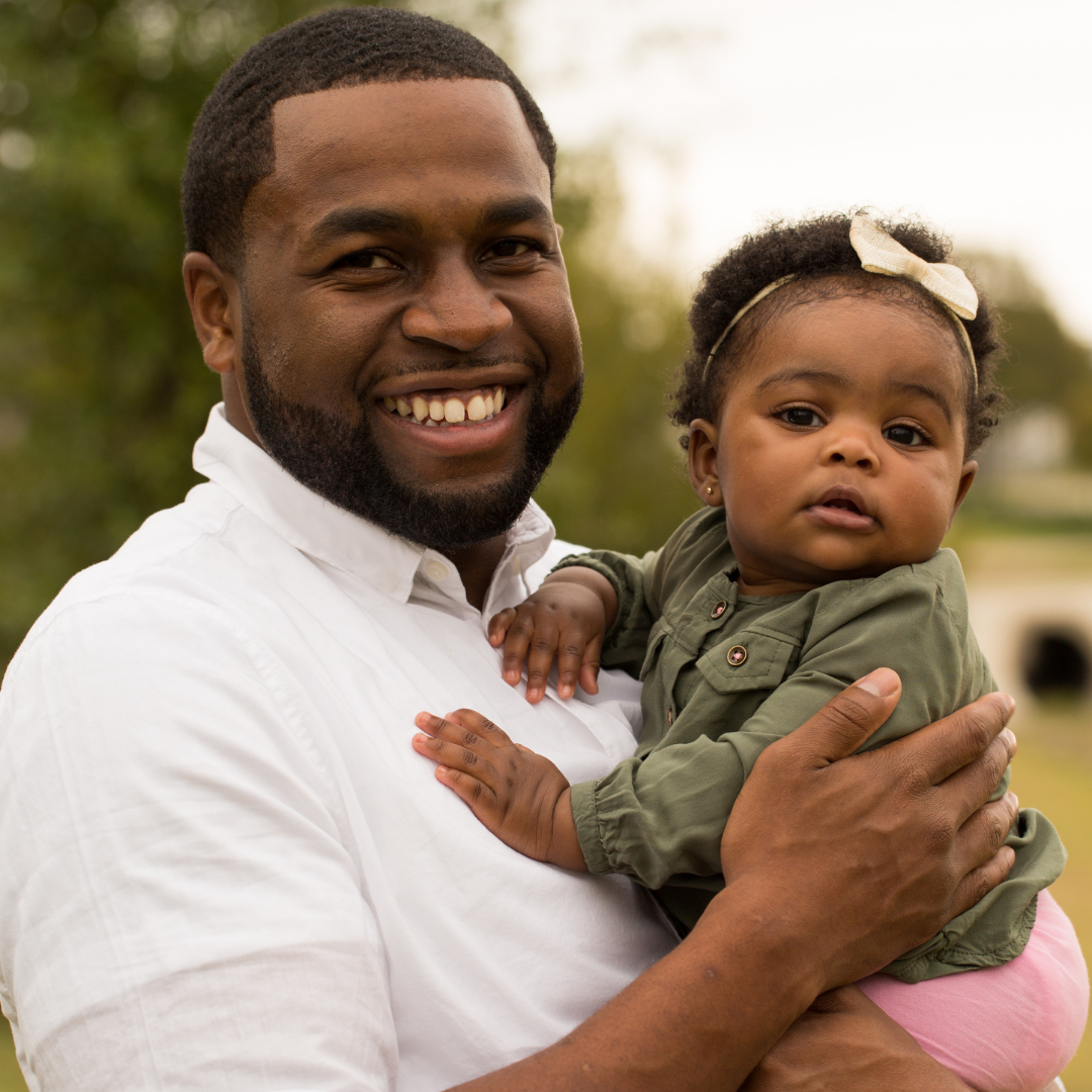 Smiling Black man holds a baby girl outdoors; both look at the camera.