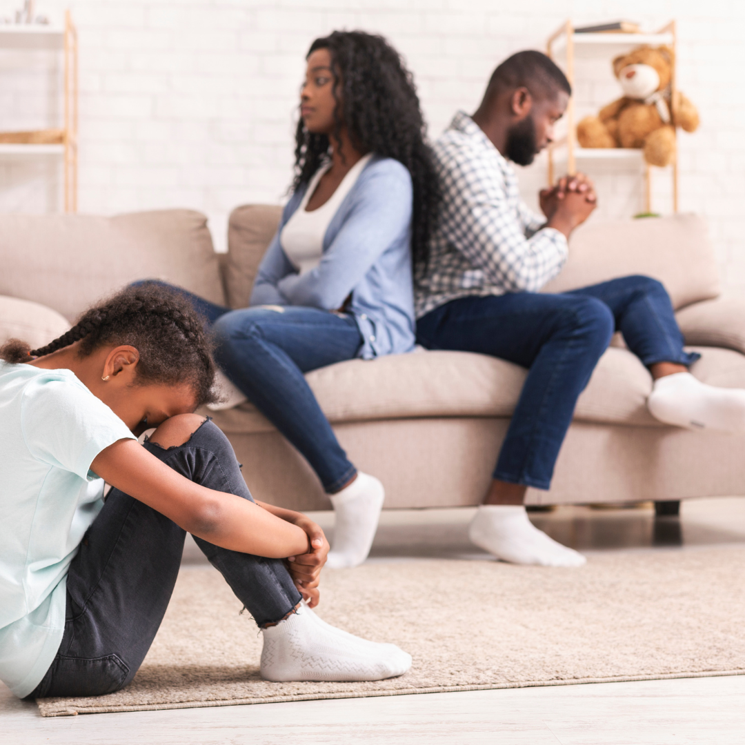 A sad girl sits on the floor, her parents sit on the couch behind her, turned away from each other.