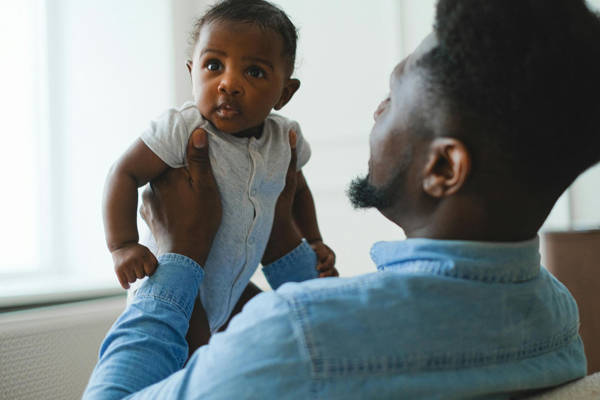 An adult holds a baby up in front of them, looking at the child with a gentle expression in a bright room.