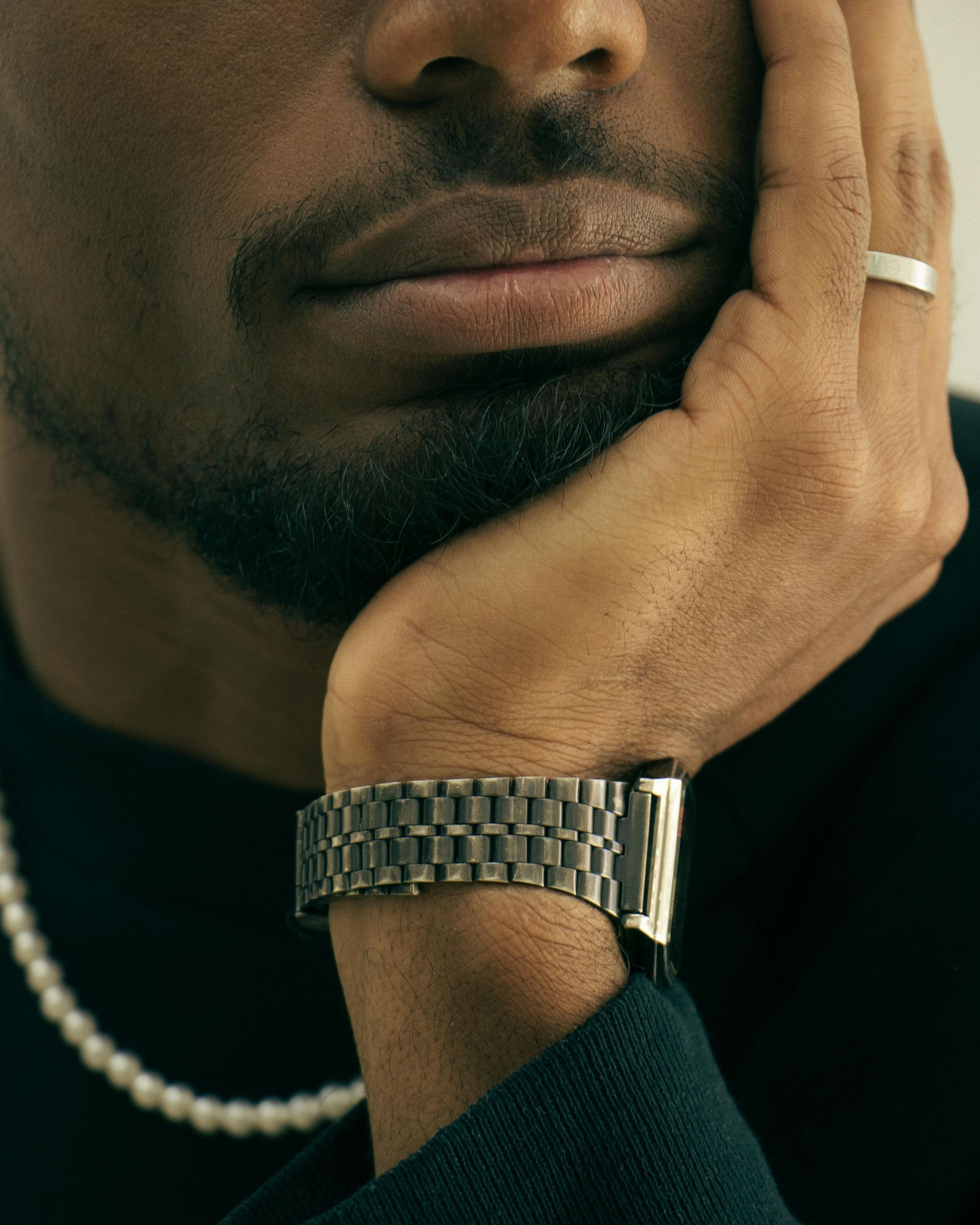 Close-up of a hand resting on a chin, featuring a metal link watch band, a simple band ring, and a pearl necklace.