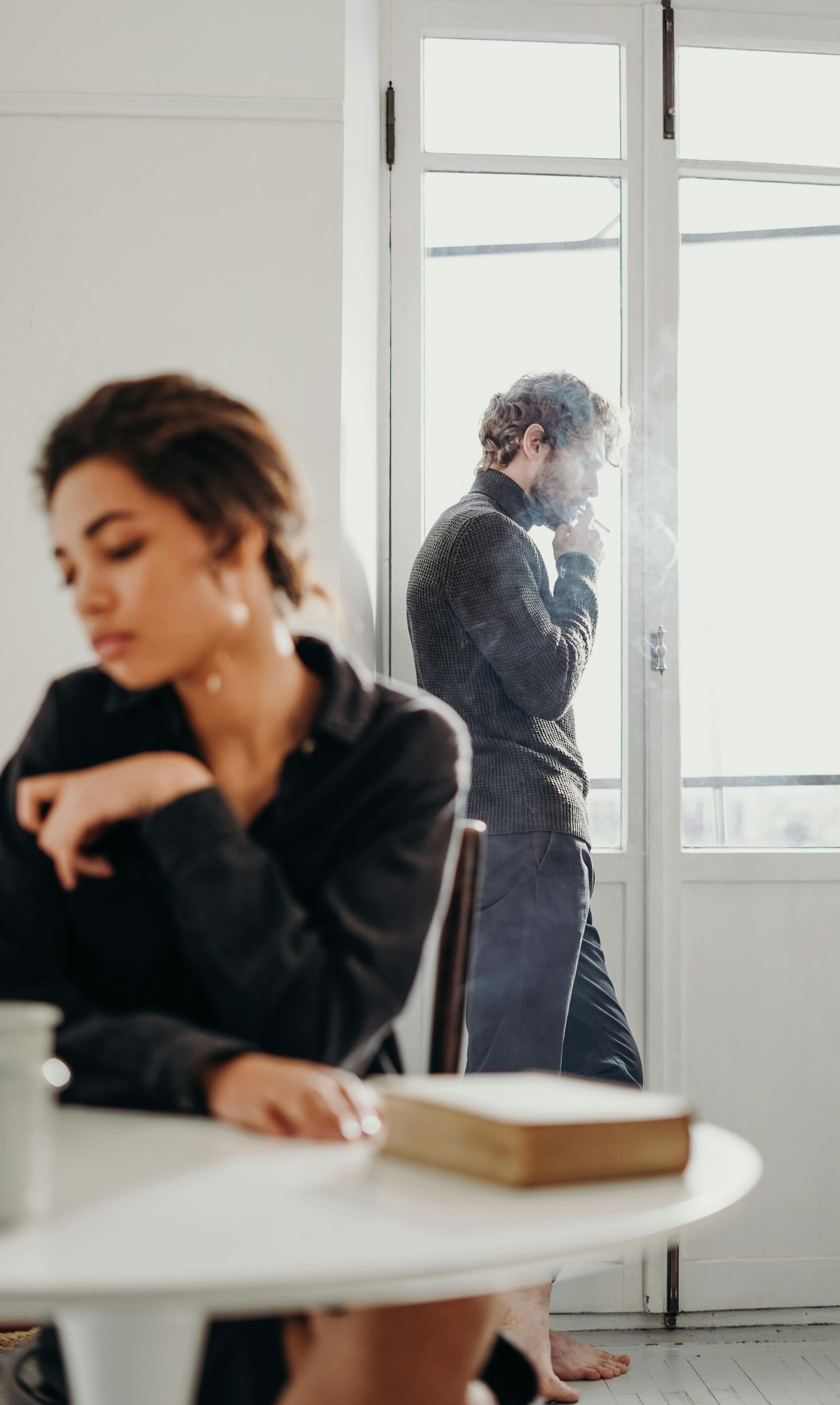 Woman at a table, looking down. Man in the background by a bright window, thoughtful.