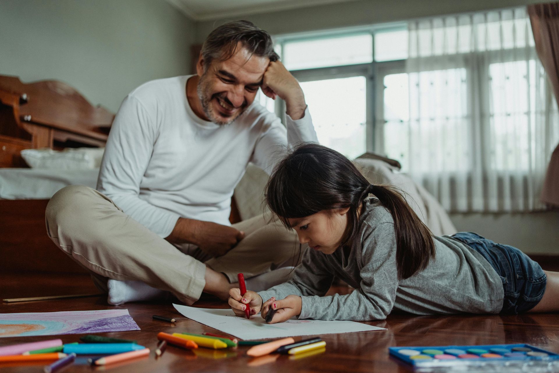 Man smiles, watching a girl draw with crayons on the floor in a room with a window.