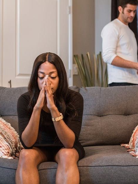 Woman with hands clasped, looks distraught on a couch. Man in background, looks away.