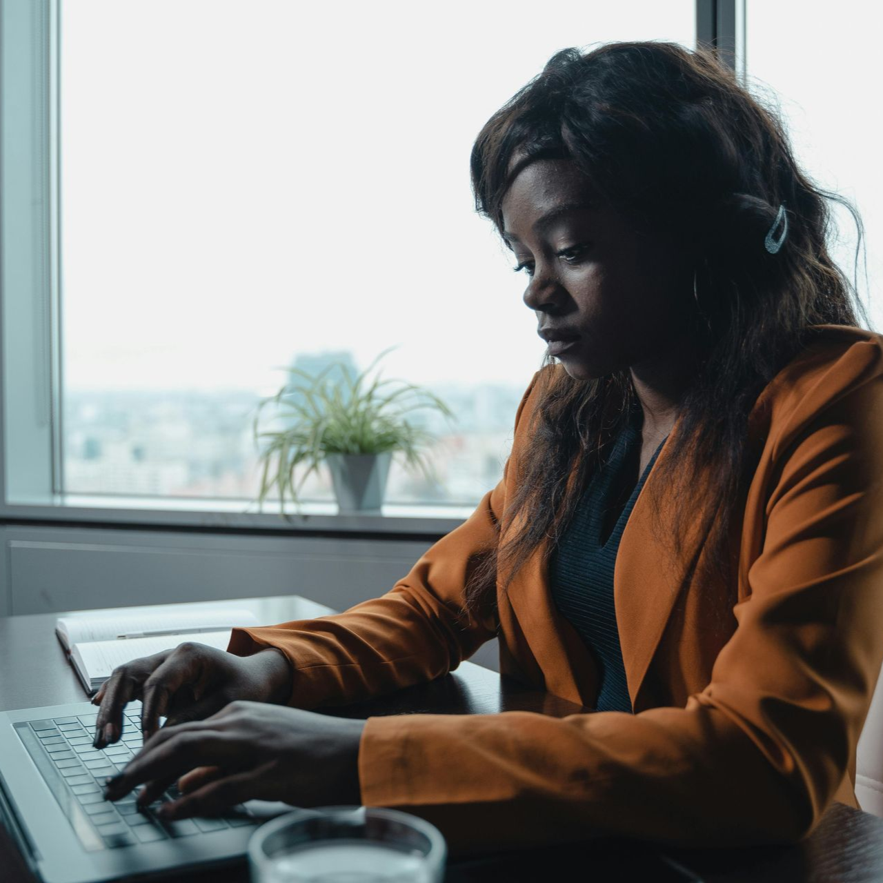 Woman in orange blazer types on a laptop at a table near a window.