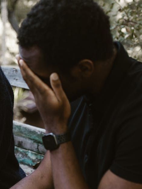 Black man, face in hands, appears to cry, sitting outdoors on a bench; he wears a black shirt and watch.