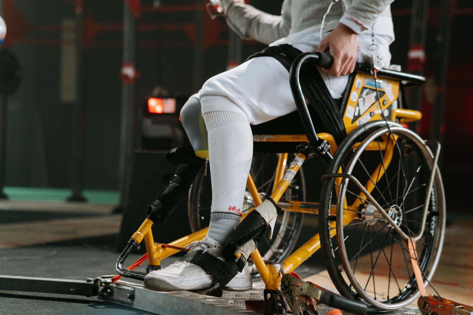 A wheelchair fencer in white protective gear competes, secured in a yellow specialized frame on a sports floor.