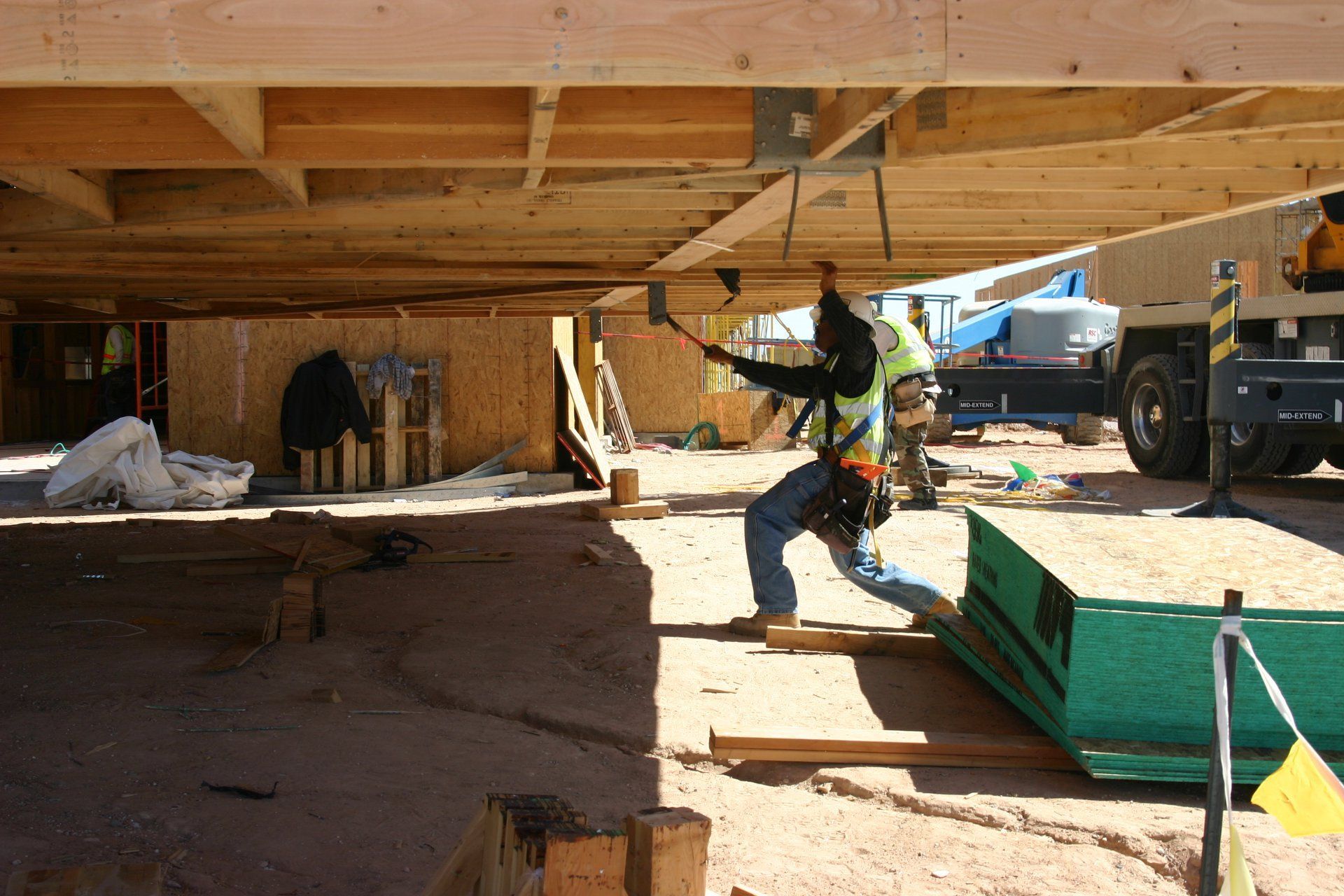Construction worker under a house, handling a hose near green equipment and a truck in the background
