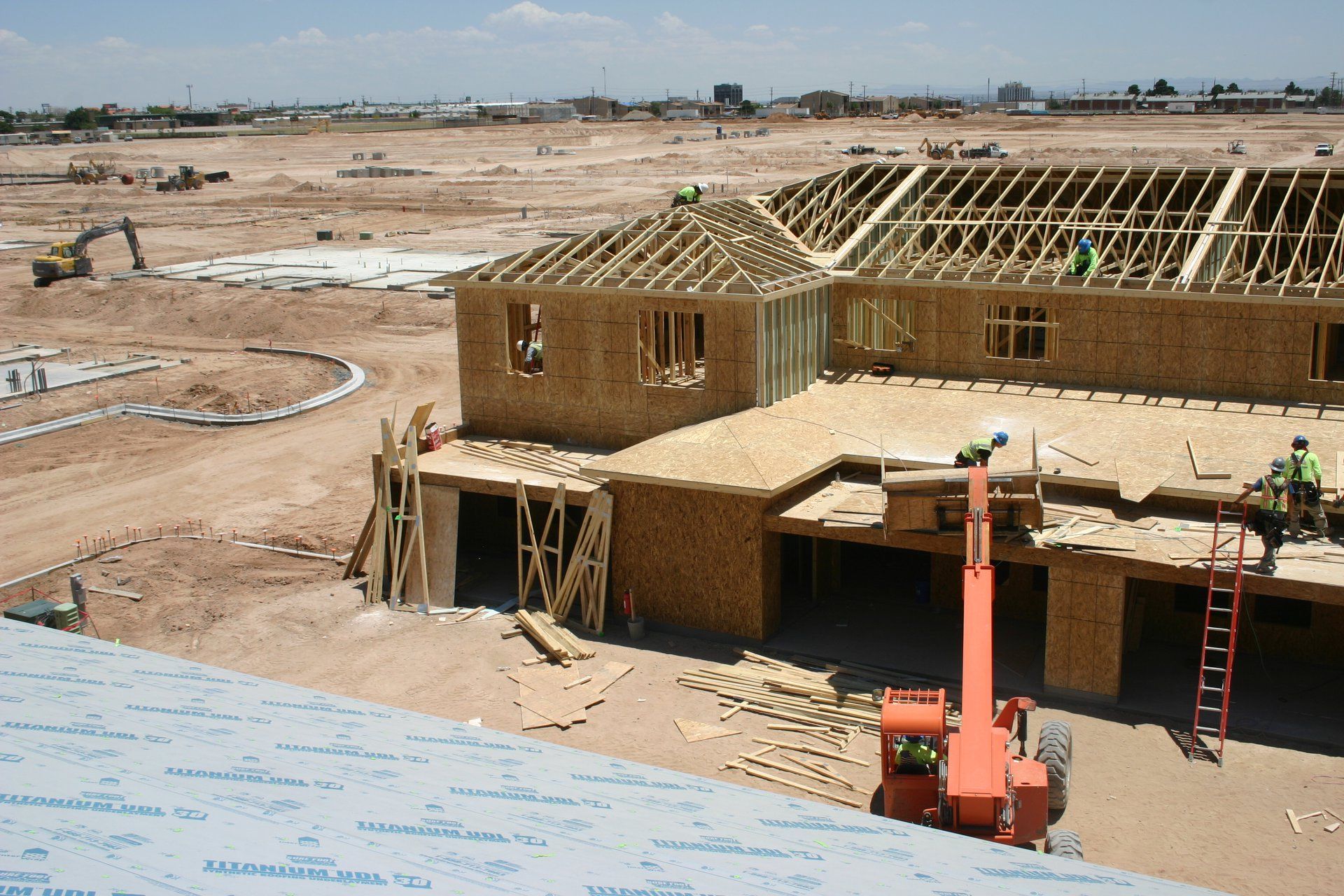 House under construction in a sandy lot, with exposed roof framing and workers on site.