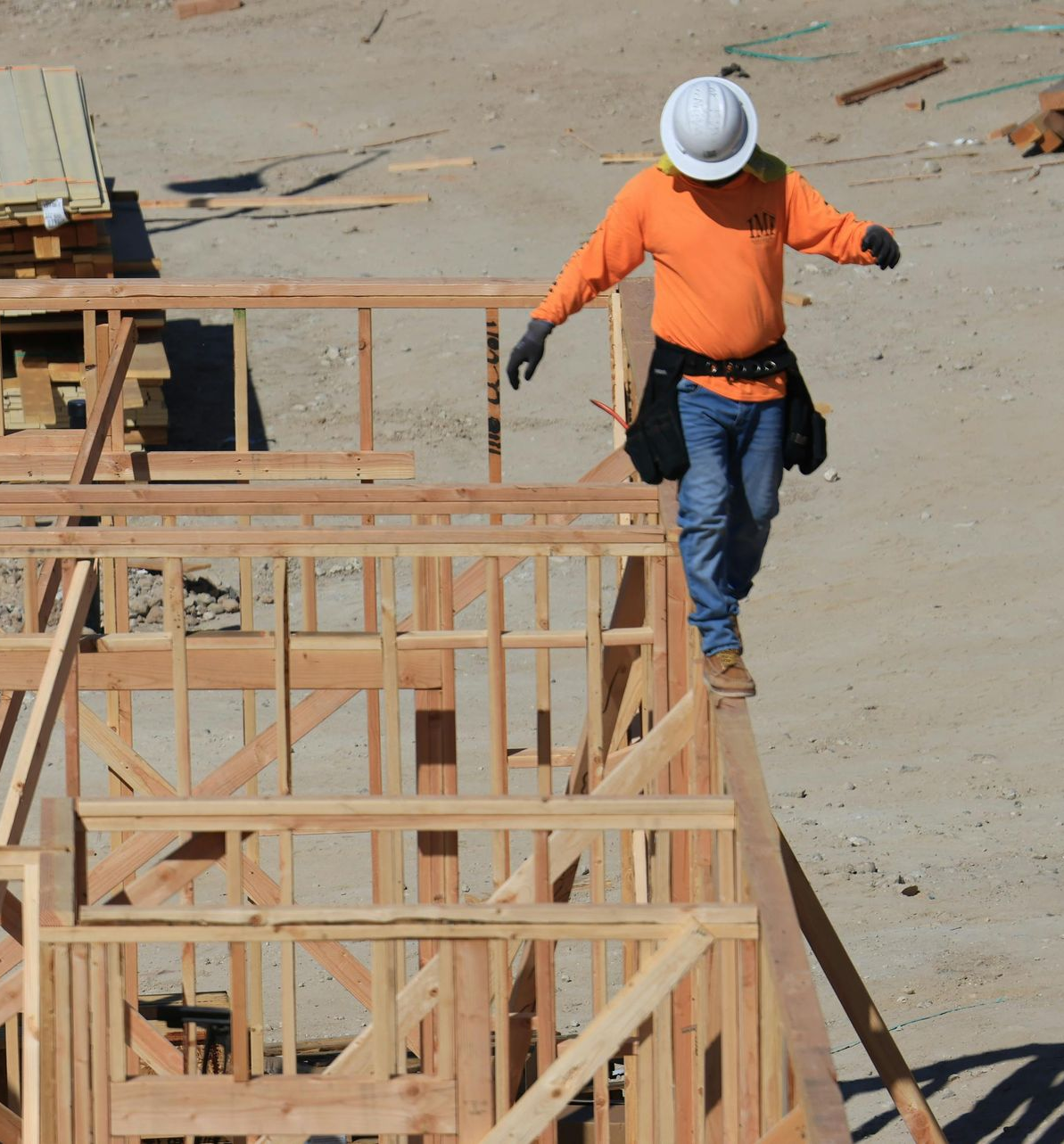 Construction worker in orange shirt and hard hat walking along wooden framing at a building site