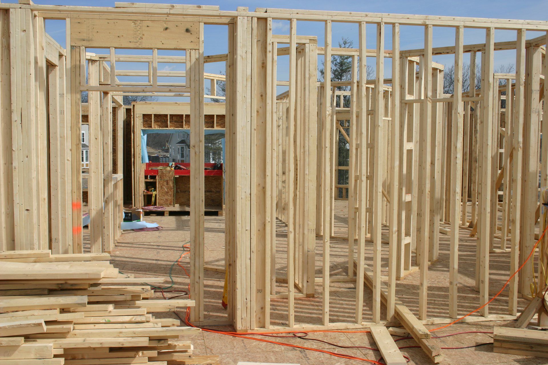 Wood-framed house under construction with stacked lumber and a red truck visible through the studs