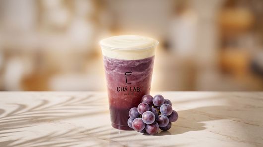 Purple grape smoothie in a clear cup with a creamy foam top, shown with fresh grapes on a wooden surface.