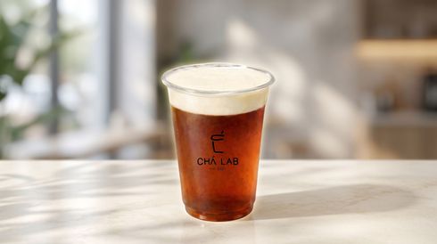 A clear plastic cup of iced coffee with foam, standing on a sunlit table by a window.