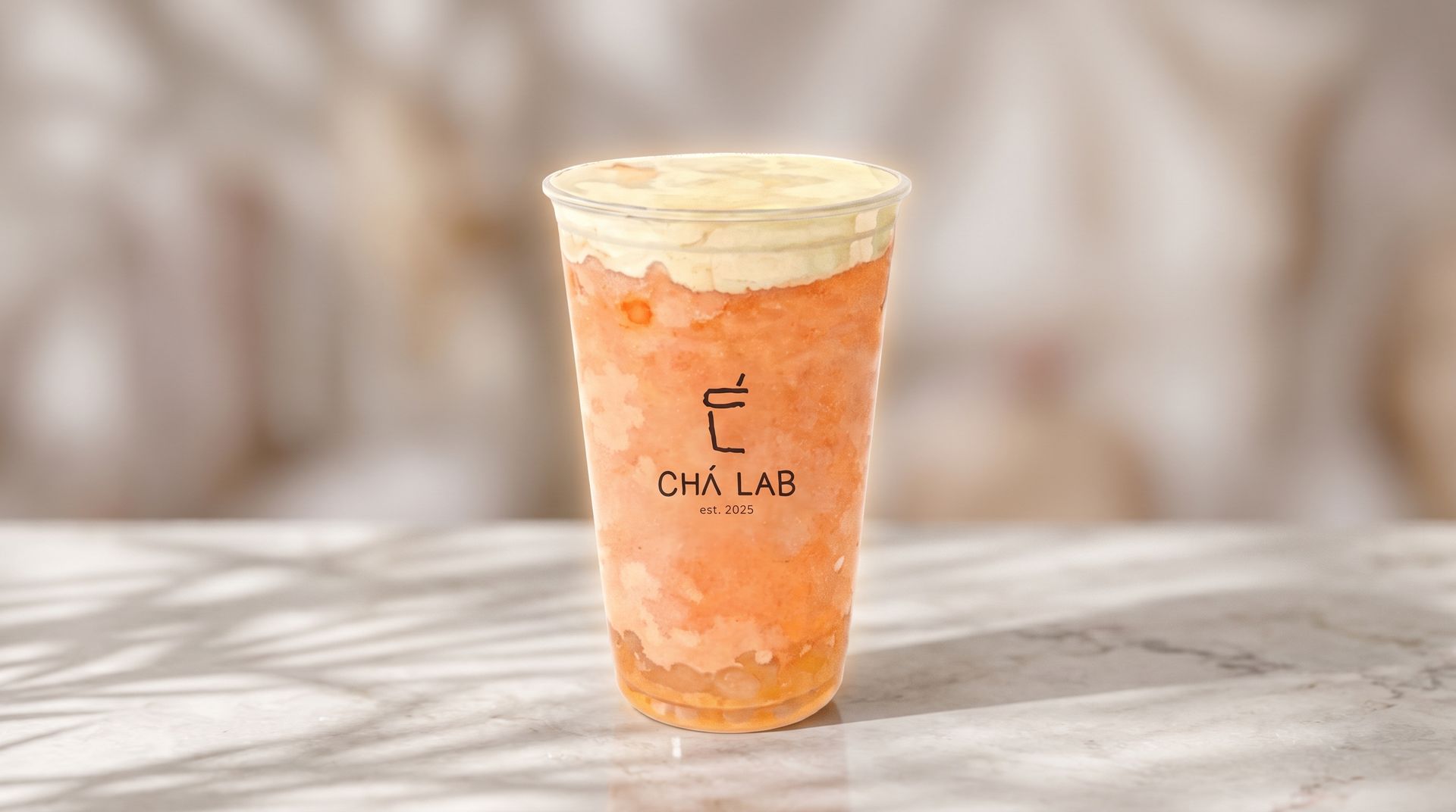 Clear cup of orange iced drink topped with cream on a marble surface, with a softly blurred background