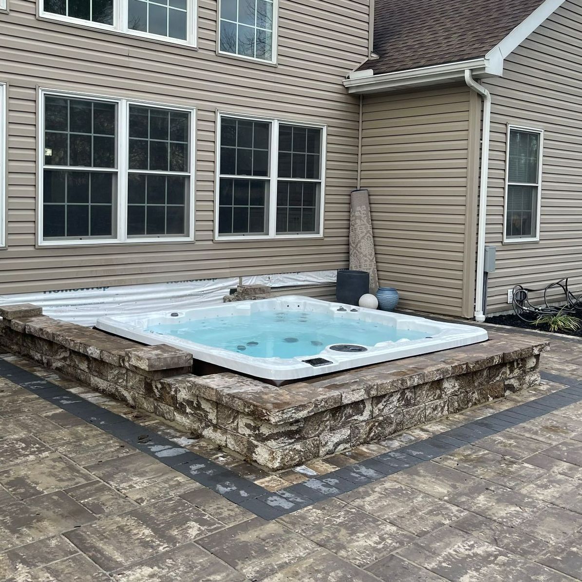 Hot tub on a stone patio next to a two-story house with multiple windows.