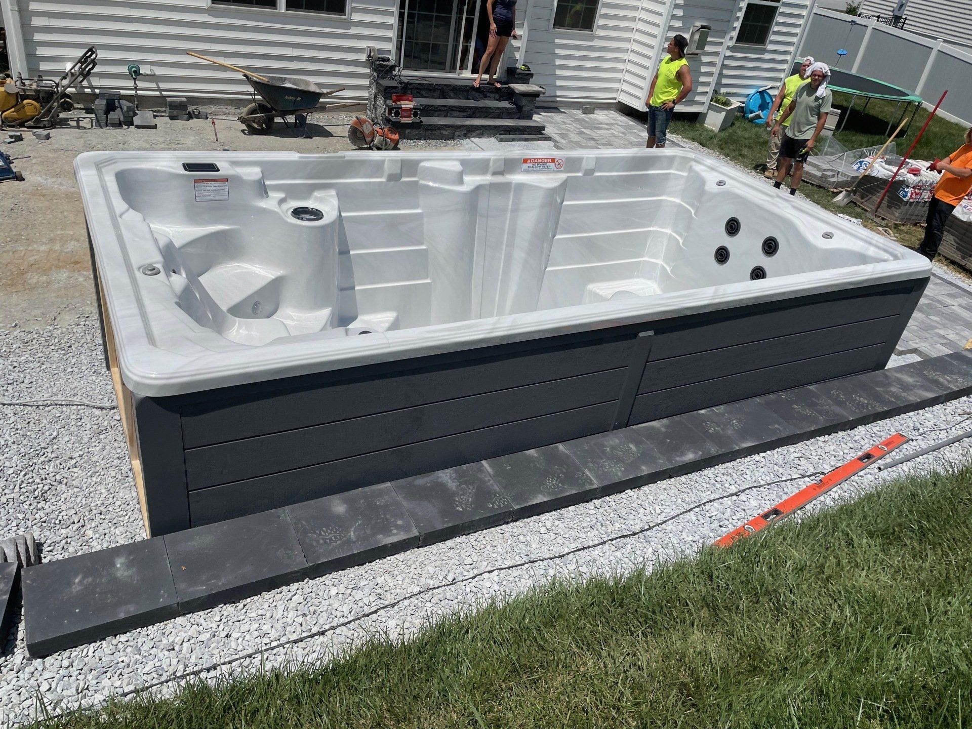 A rectangular hot tub installation outdoors. Gray exterior, white interior, gravel base, and workers nearby.