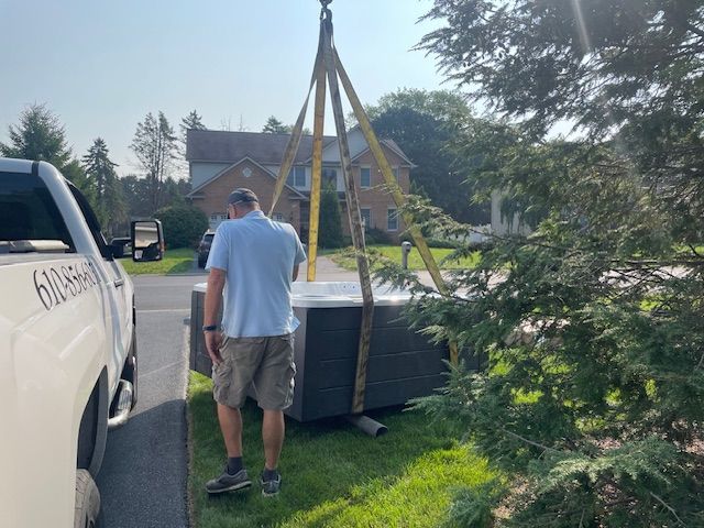 Man supervises a crane lifting a hot tub into a yard near a truck and house.