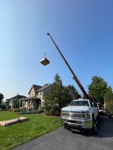 A crane lifting a large, square object near a white truck parked in a driveway in front of a house on a sunny day.
