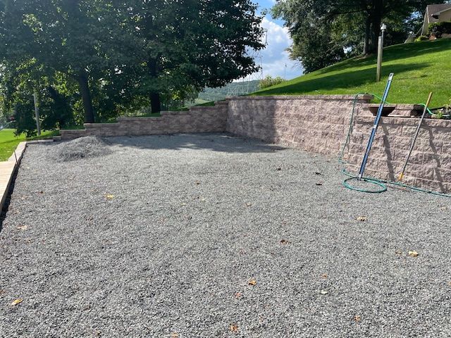 A gravel patio with retaining walls, a hose, and a grassy hill in the background.