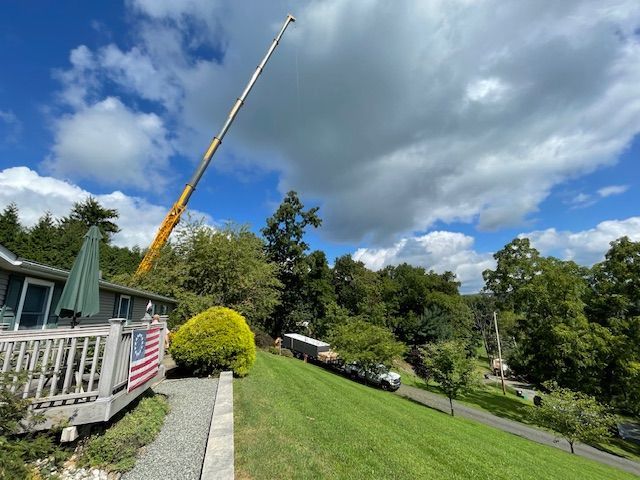 Crane lifting something. Green grass, trees, cloudy sky. House with deck, American flag.