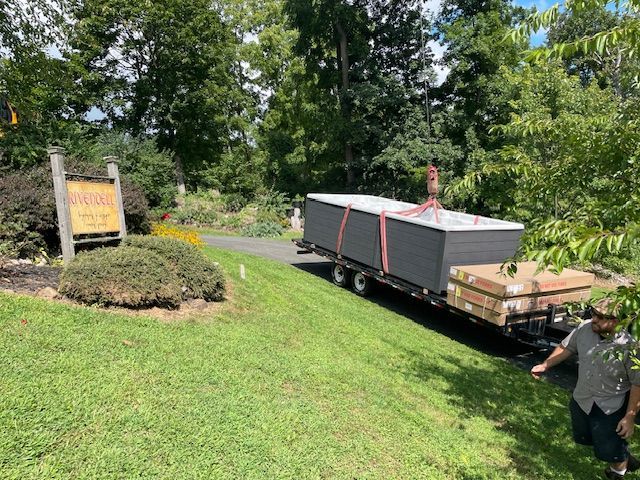 A trailer carrying a large, rectangular structure uphill toward a sign and trees, with a person walking alongside.