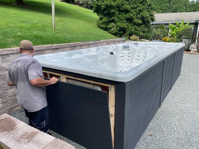 Man installing dark gray panel on a large hot tub in a backyard setting.