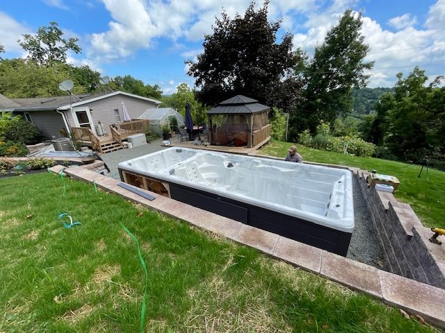 A swim spa sits in a landscaped yard. A man works nearby. A house and gazebo are in the background.