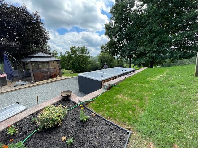 A gray hot tub sits on a raised platform next to a gravel path and a grassy hill, outdoors.