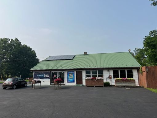 Small, white building with green roof and solar panels, storefront with signs, flowers, and a parked car.