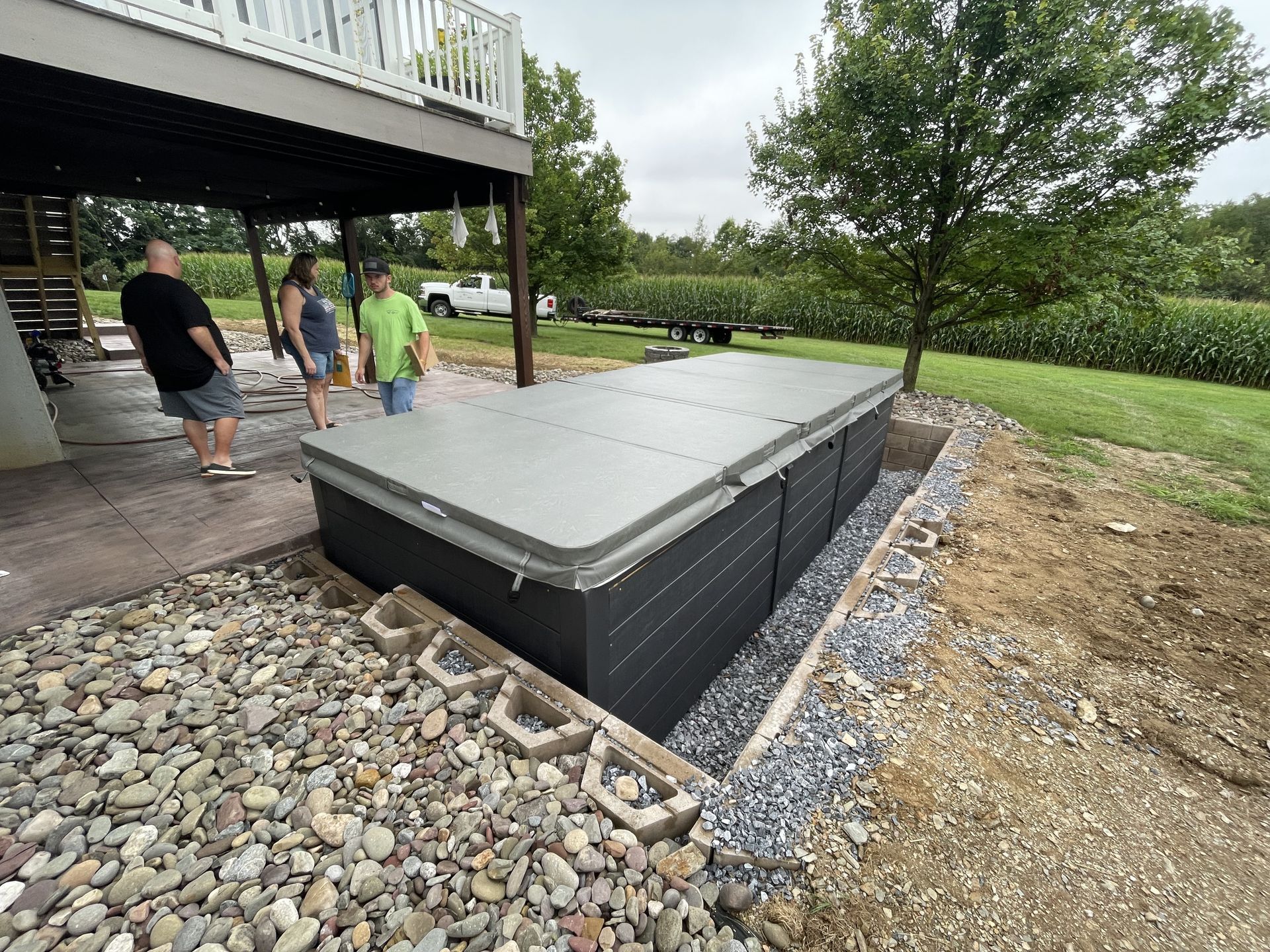 A rectangular, covered hot tub next to a house. People stand nearby.