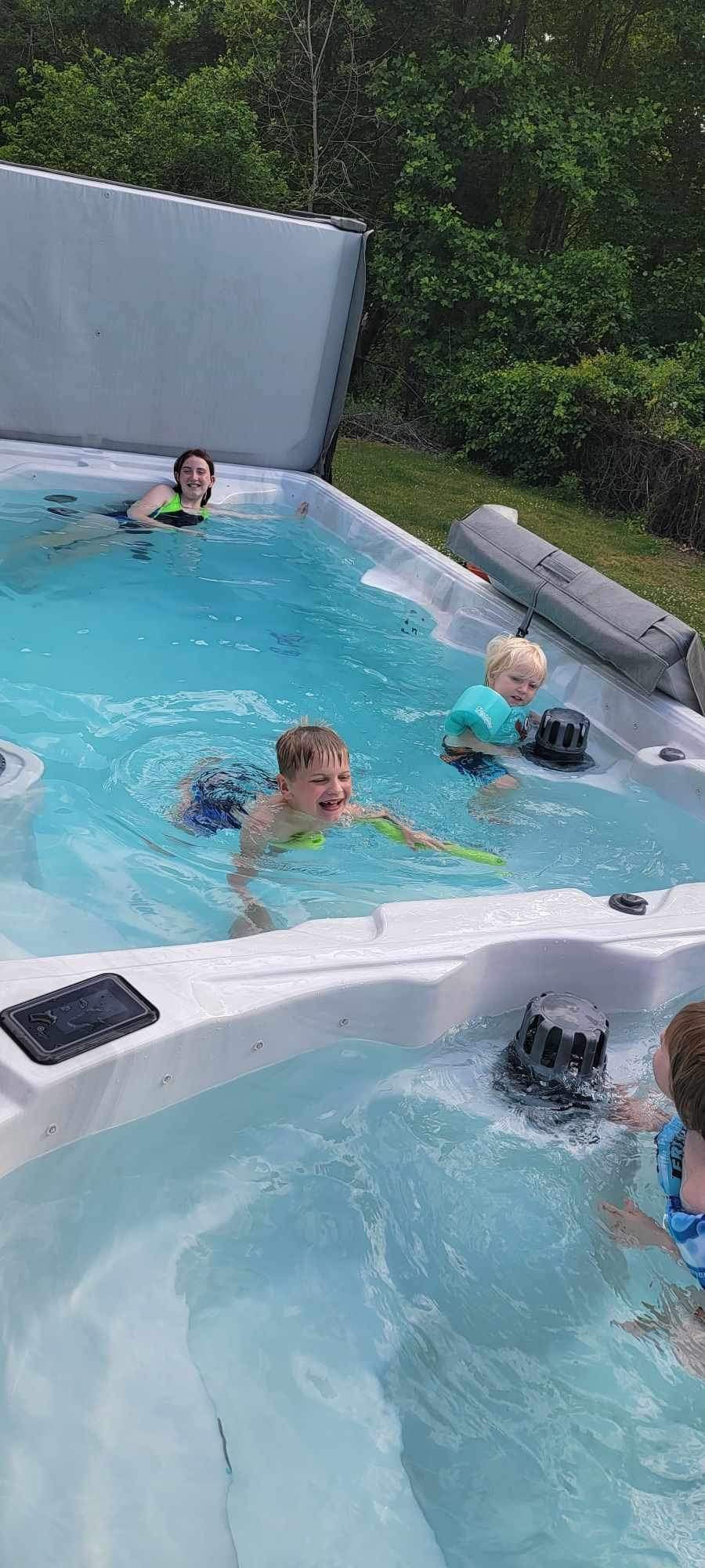 Children playing in a hot tub, surrounded by trees. One child swims while others float.