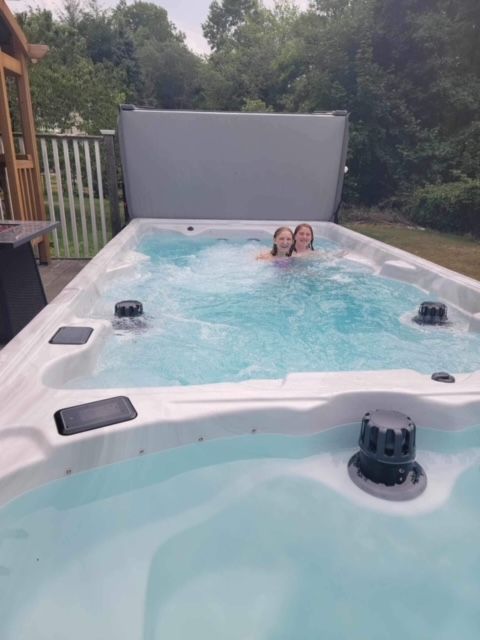 Two people in a long, rectangular swim spa. The spa is outdoors, near trees and a deck.