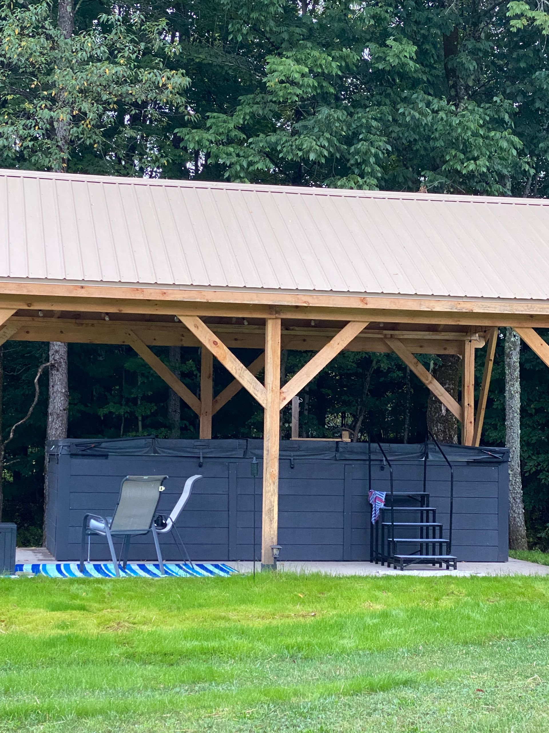 Open-sided wooden shelter with patio furniture on a grassy lawn, backed by trees.