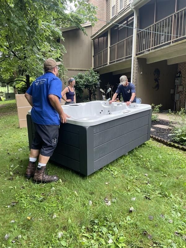 Three people moving a rectangular hot tub on grass near a building. One man wears a blue shirt.