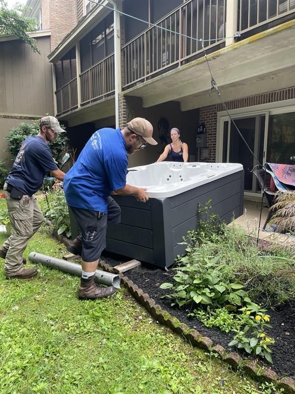 Two men lifting a hot tub in a yard, a woman watches from a patio.