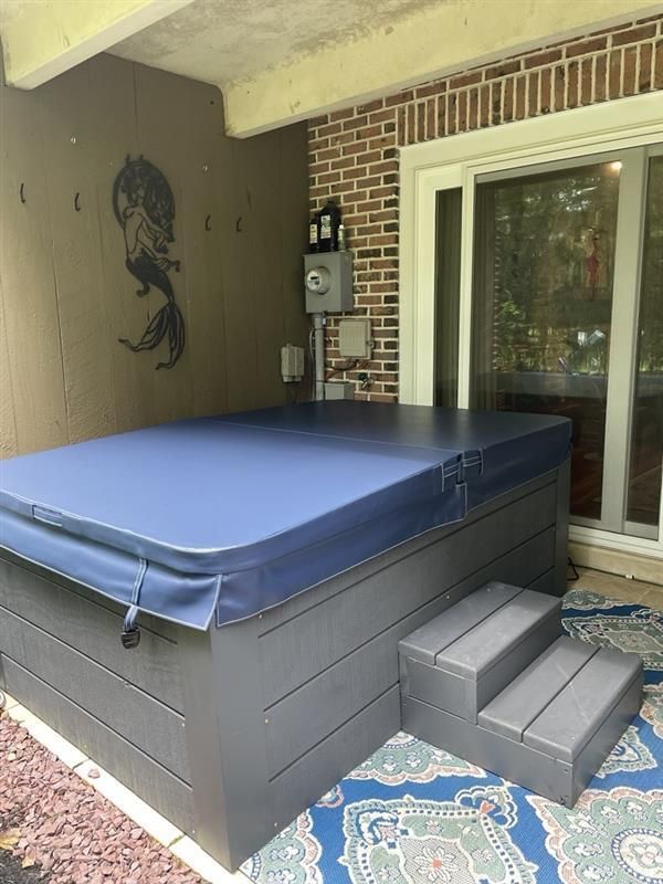Hot tub with blue cover and steps, on a patio, under a roof. Gray tub, brick wall.