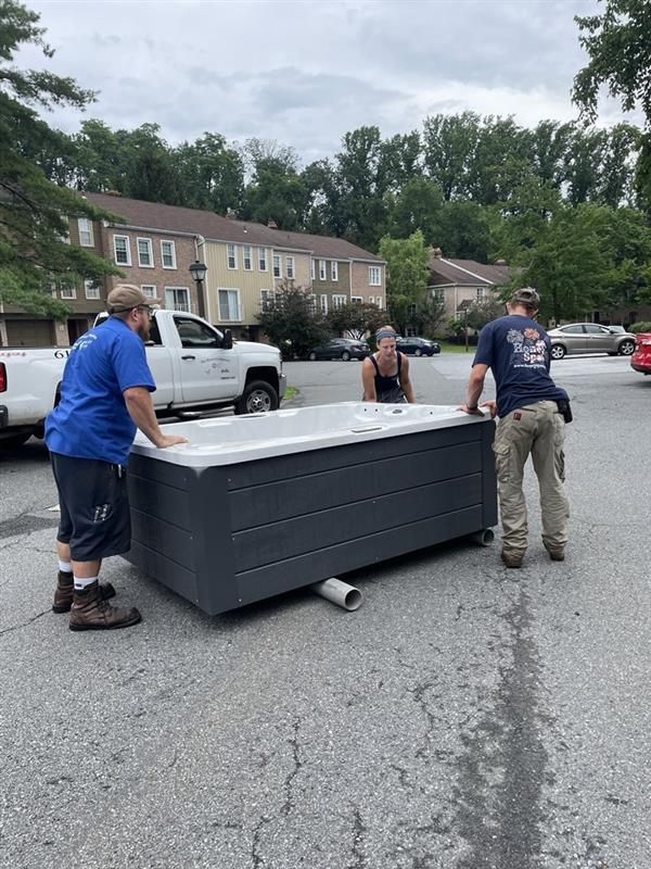 Three people moving a dark gray hot tub on wheels down a paved driveway, residential setting.