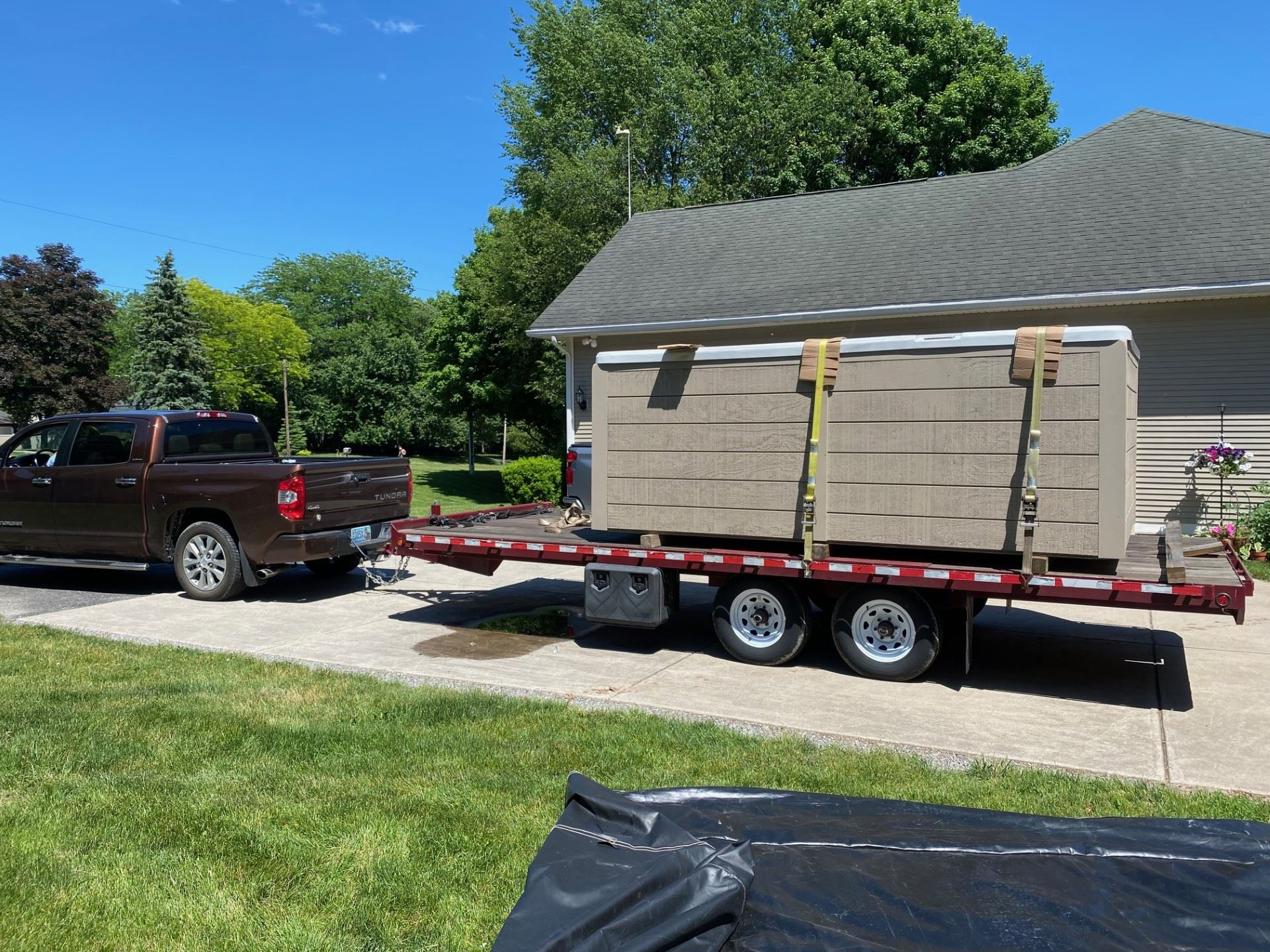 Brown pickup truck towing a trailer with a large tan rectangular object in a residential driveway on a sunny day.