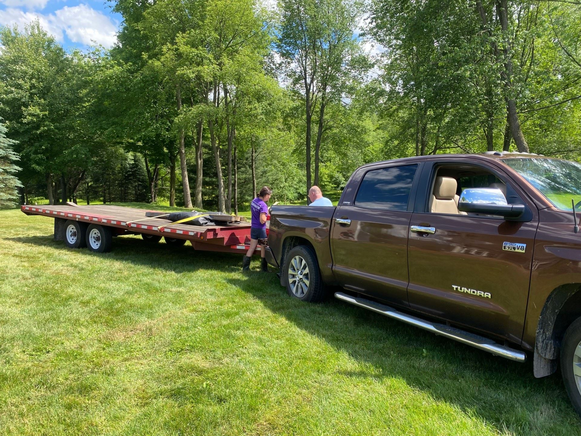 Brown truck towing a red flatbed trailer on a grassy field, two people nearby, trees in background.