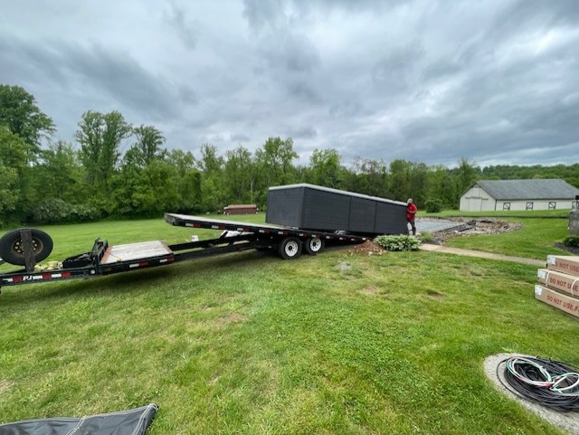 Black trailer carrying a dark rectangular structure on a grassy lawn, with person nearby.